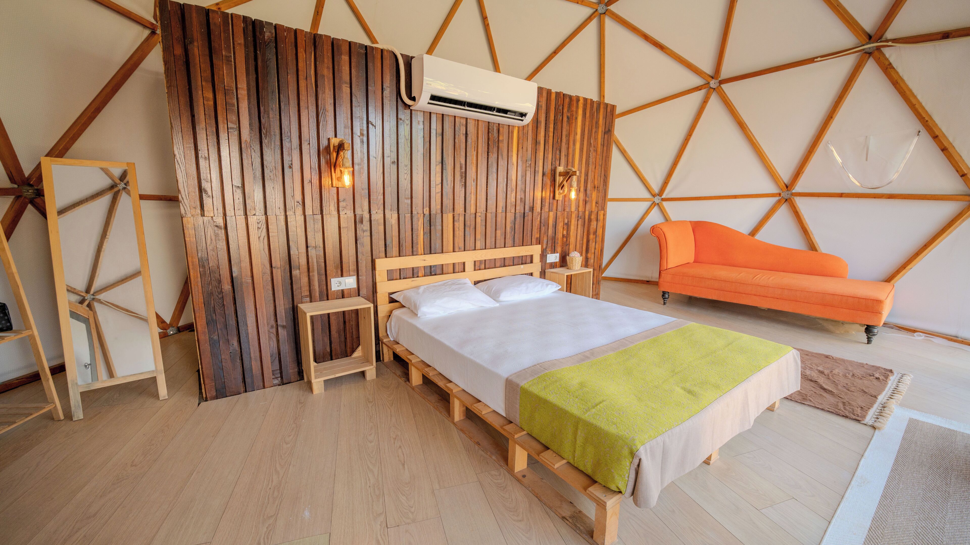Quarto com parede de ripas de madeira, cama com colchão branco e manta verde, sofá laranja e escada de madeira.
