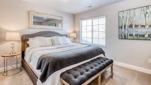 A bedroom with a large bed, a bench at the foot, a window with blinds, a mirror above the bed, and a painting on the wall.