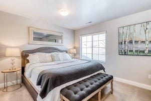 A bedroom with a large bed, a bench at the foot, a window with blinds, a mirror above the bed, and a painting on the wall.