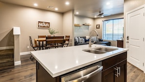 A modern kitchen with a white countertop, stainless steel sink, and built-in dishwasher. There is a dining area with a wooden table and chairs, and a living room with a sofa and a ceiling fan. - Sand Cove 3 Bedroom Townhouse (Washington)