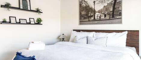 A neatly arranged bedroom with a bed, pillows, a headboard, a ceiling fan, a wall-mounted shelf with plants, framed pictures, and a large black-and-white cityscape painting.