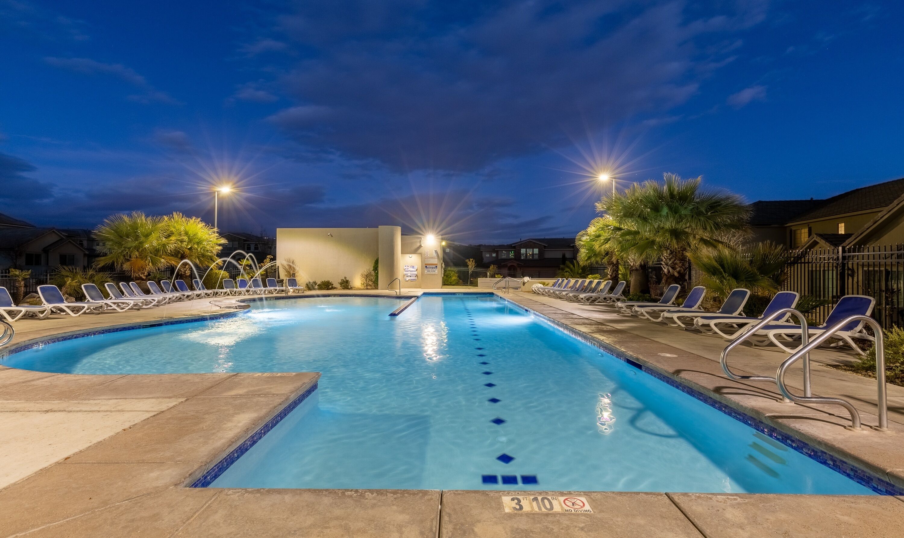 A swimming pool with lounge chairs and a no diving sign, surrounded by palm trees and houses.