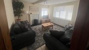 A living room with two black sofas, a wooden coffee table, a fireplace, a potted plant, and a large window with blinds. - Blyton 2-bed Cottage, Parking, 30m to Lincoln (Gainsborough)