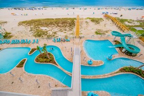 Un parque acuático con múltiples piscinas y toboganes, una zona de playa con sombrillas y un paseo de madera que conduce a las piscinas.