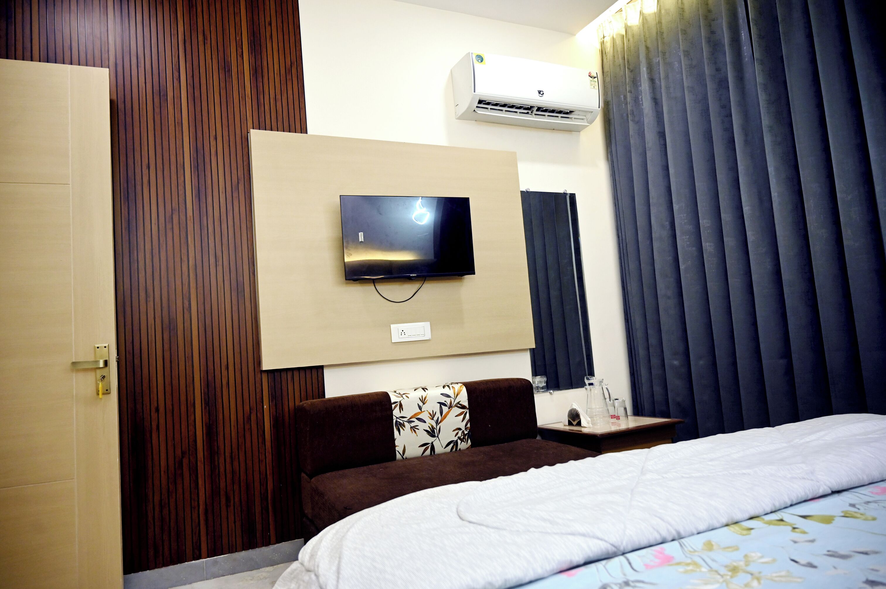 Een hotelkamer met een bed, een aan de muur bevestigde televisie, een houten deur en een raam met donkere gordijnen.