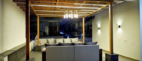A modern interior with a sofa set under a wooden pergola, a ceiling with linear lights, and a view of a building outside.