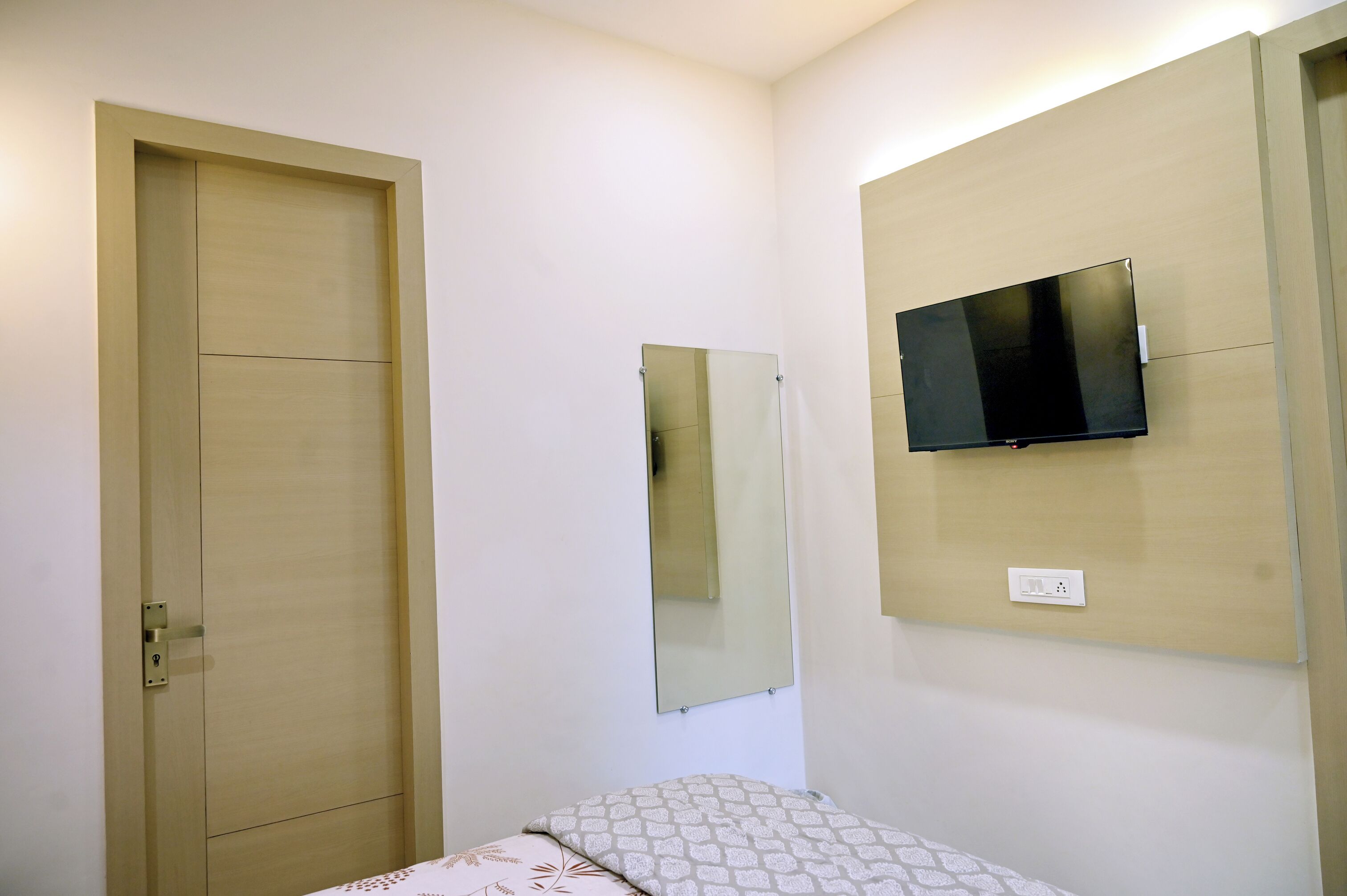 A bedroom with a wooden wardrobe, a bed with patterned bedding, a wall-mounted TV, and a mirror.