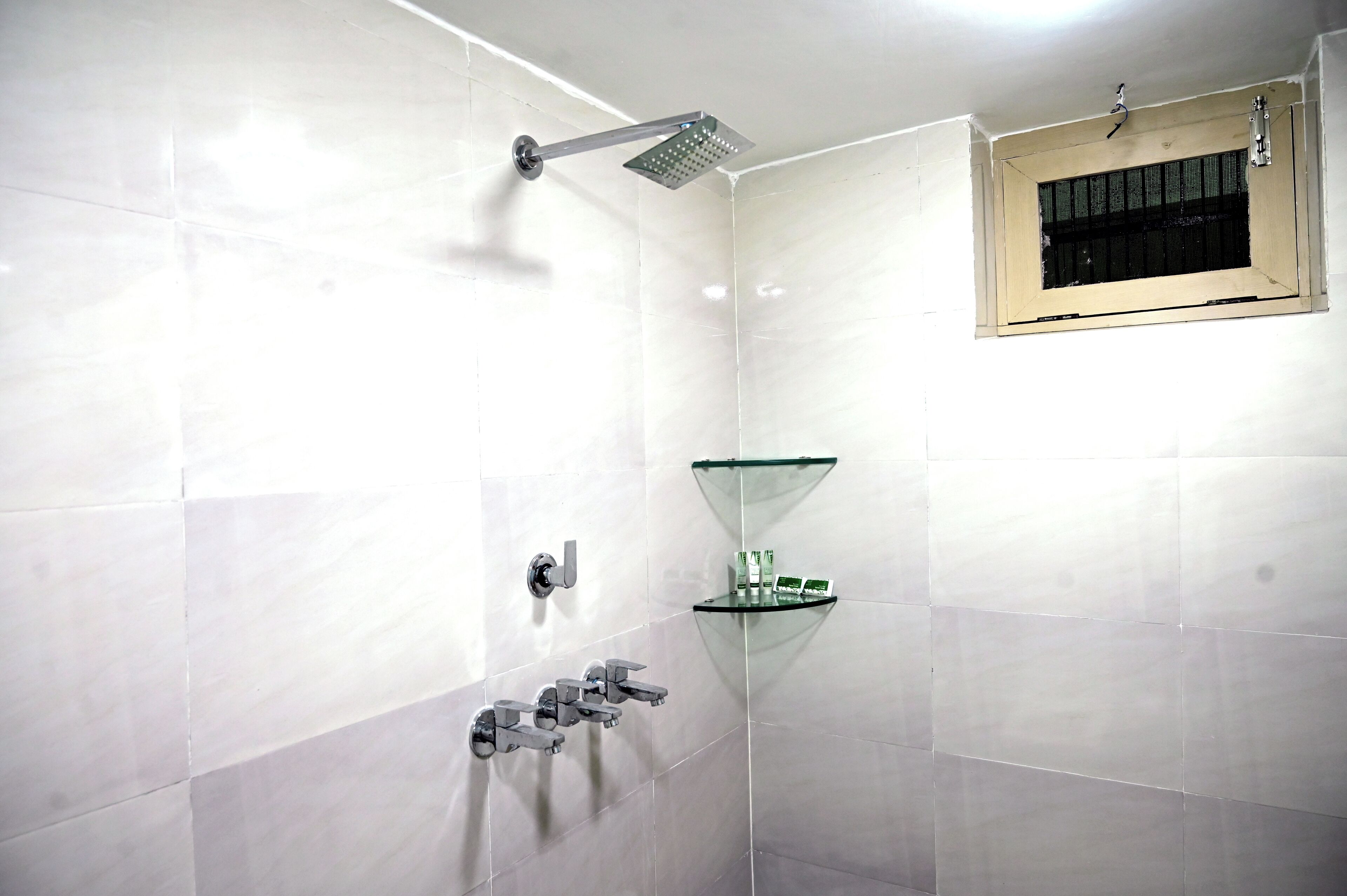 Un baño moderno con azulejos blancos, estante de vidrio, cabezal de ducha y una ventana con barrotes.