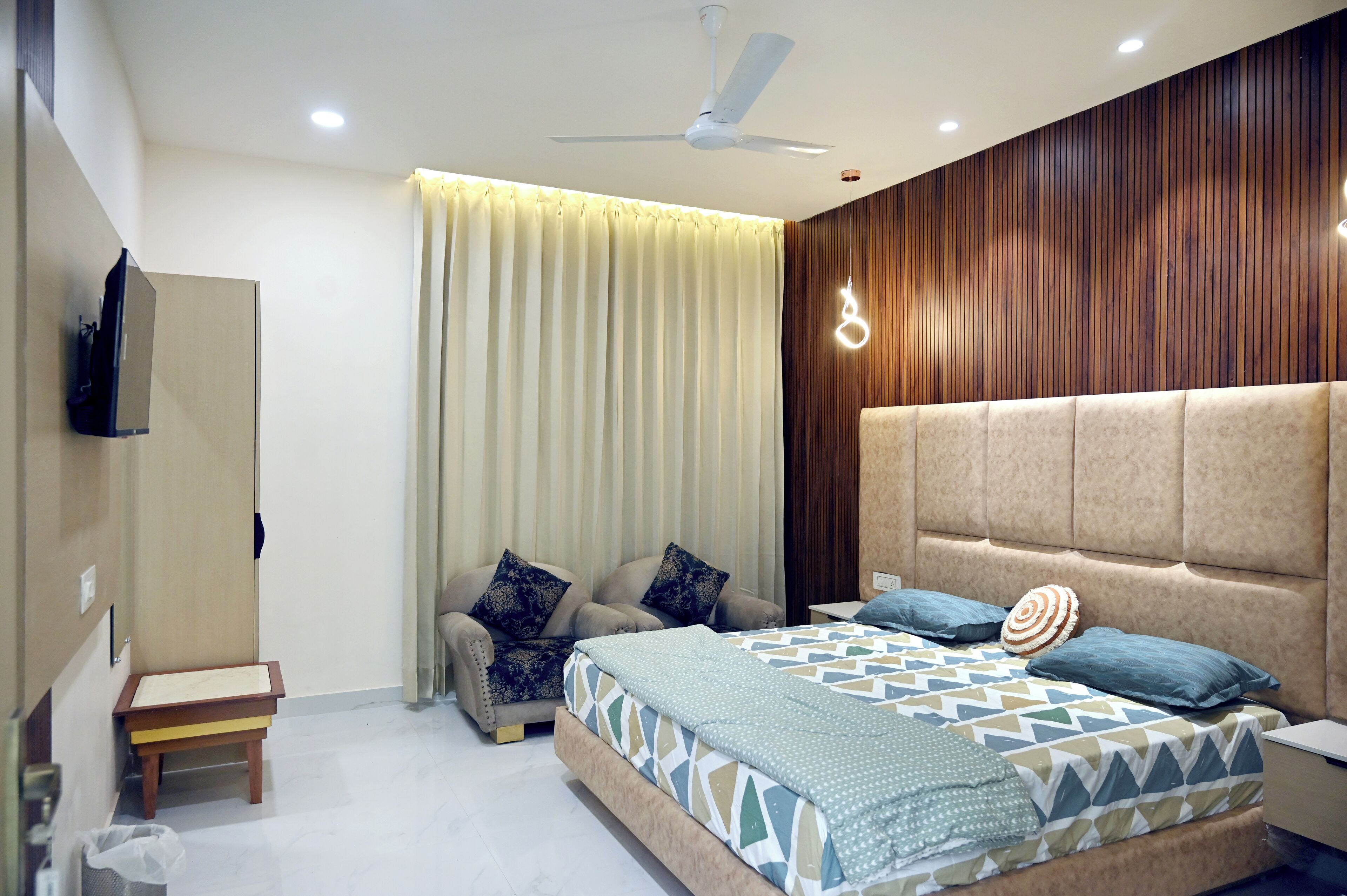 A bedroom with a bed, a sofa, a small table, a ceiling fan, and a TV mounted on the wall.