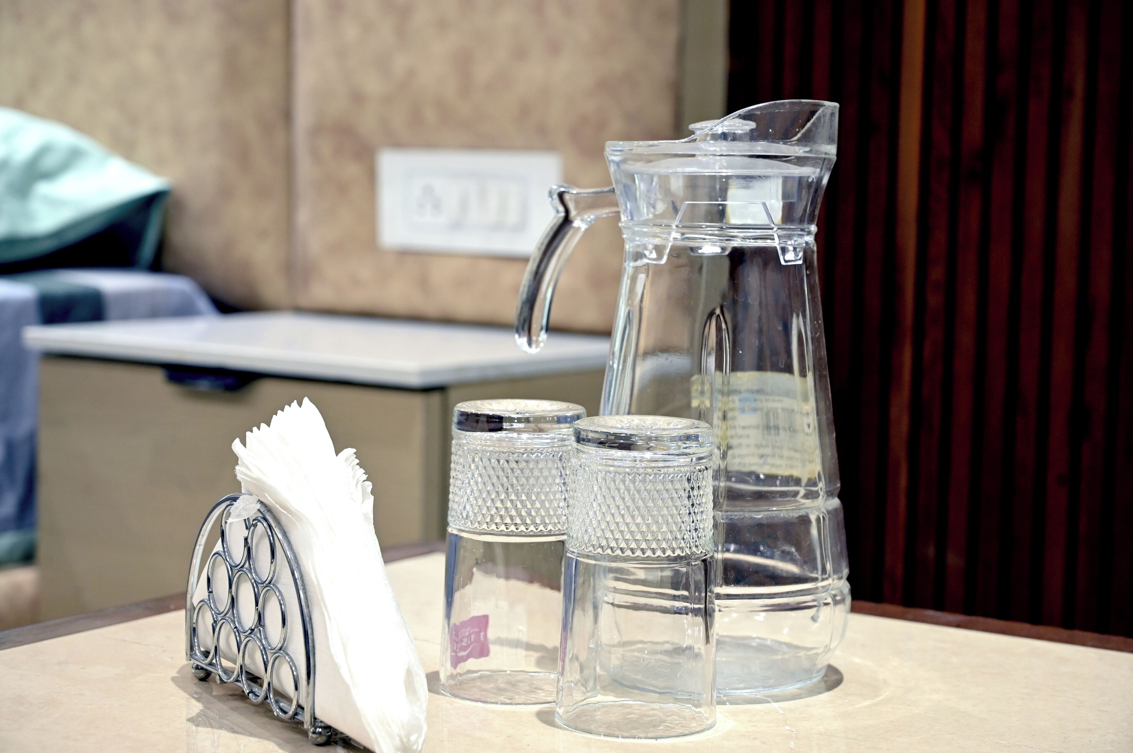 Un jarro de vidrio transparente, dos cocteleras de vidrio y un posavasos metálico sobre una mesa.