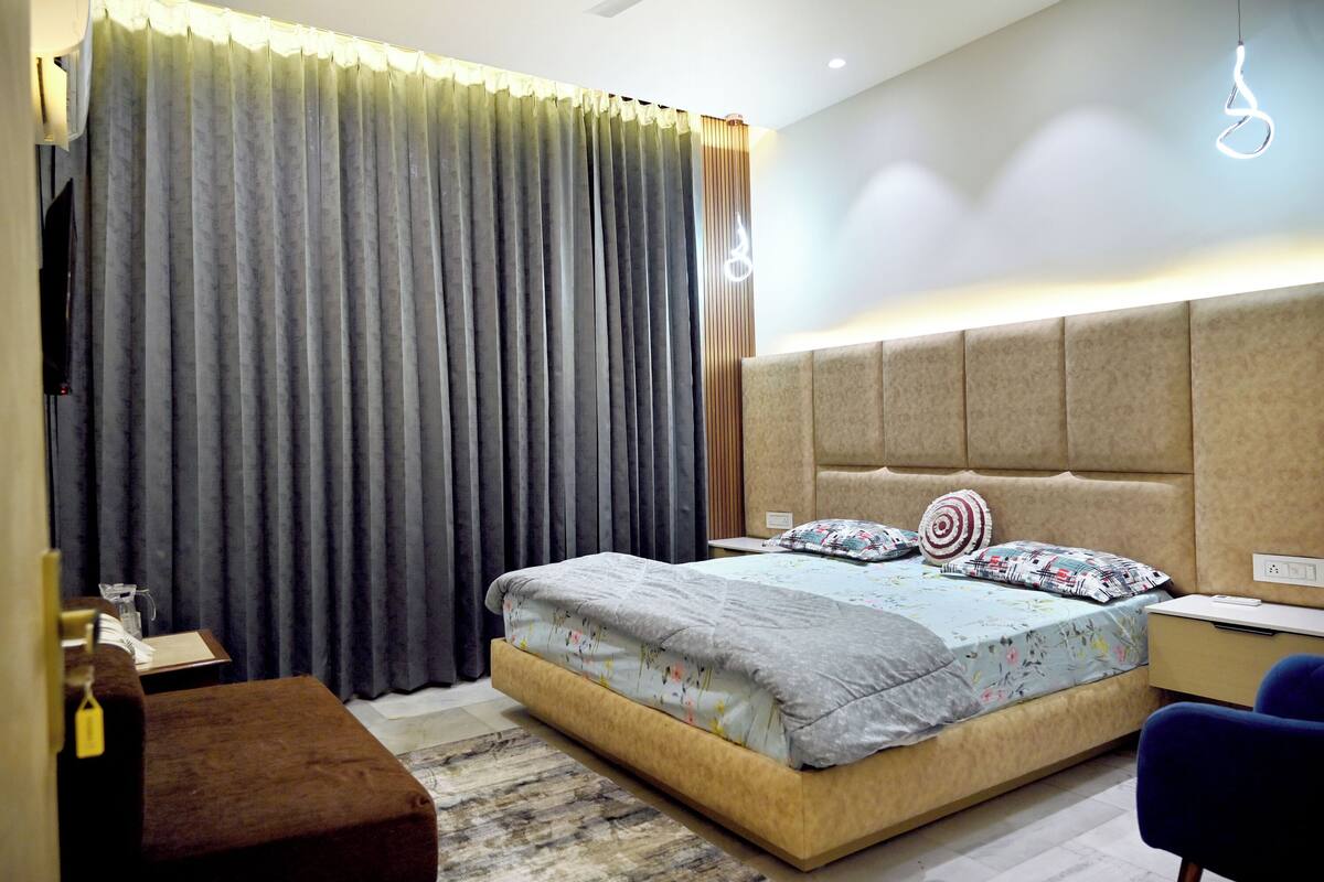 A bedroom with a bed, a desk, a chair, a TV, and a large window with curtains.