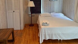 Un dormitorio con una cama, una mesita de noche, una lámpara, una ventana con persiana negra y suelo de madera.