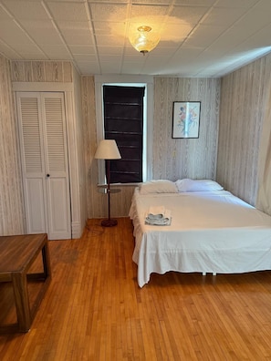 A bedroom with a bed, a nightstand, a lamp, a window with a black blind, and a wooden floor.