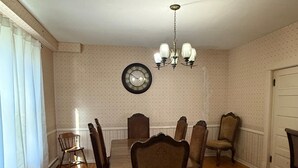 Un comedor con una mesa de madera, seis sillas, una araña, un reloj de pared y una ventana con cortinas.