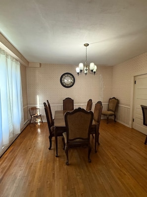 A dining room with a wooden table, six chairs, a chandelier, a wall clock, and a window with curtains.