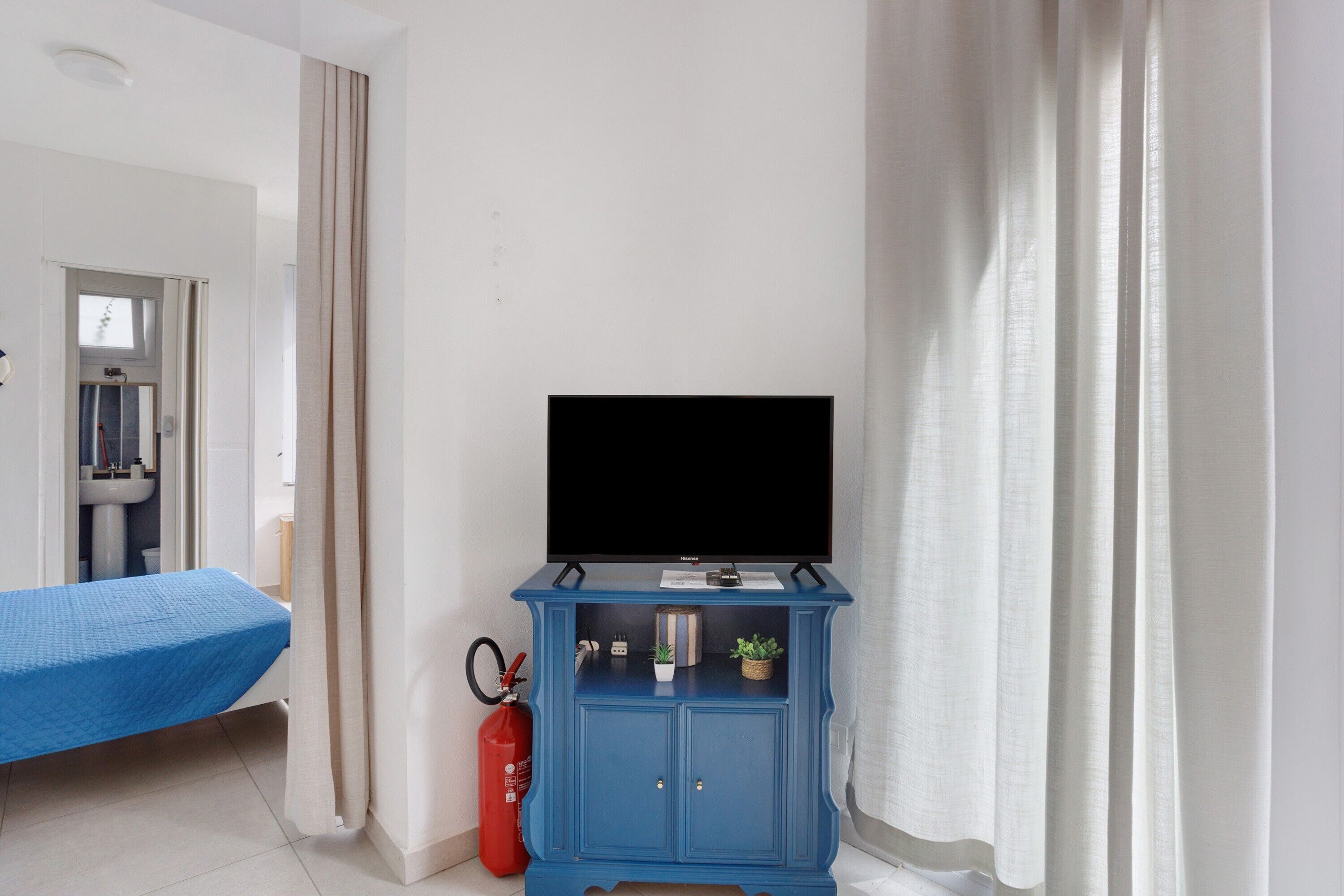 A room with a bed, a blue cabinet with a television, a fire extinguisher, and a bathroom visible through an open door.