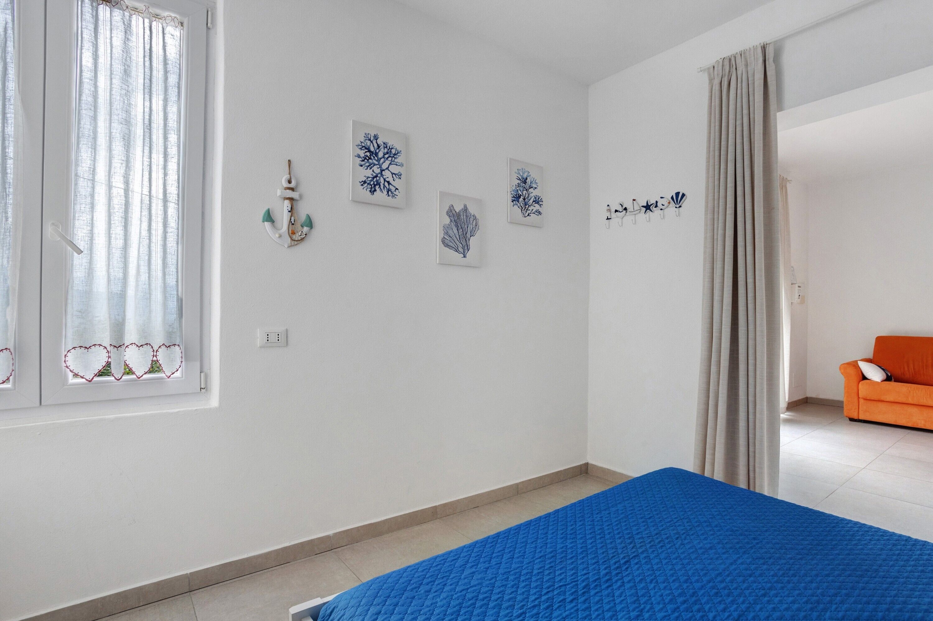 A bedroom with a bed, a window with curtains, a wall with nautical-themed artwork, and an orange sofa in the adjacent room.