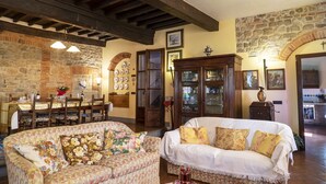 A cozy living room with a dining area, featuring a stone wall, wooden beams, and traditional furnishings.