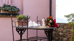 A table with a coffee pot, cups, and a bouquet of flowers on a brick patio with a pink wall and a stone wall in the background.