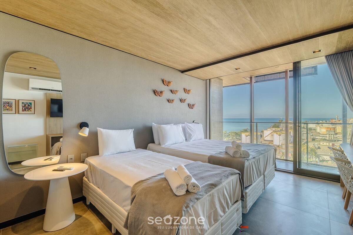 Habitación de hotel moderna con dos camas, una mesita redonda, un espejo grande y un balcón con vista a la ciudad.