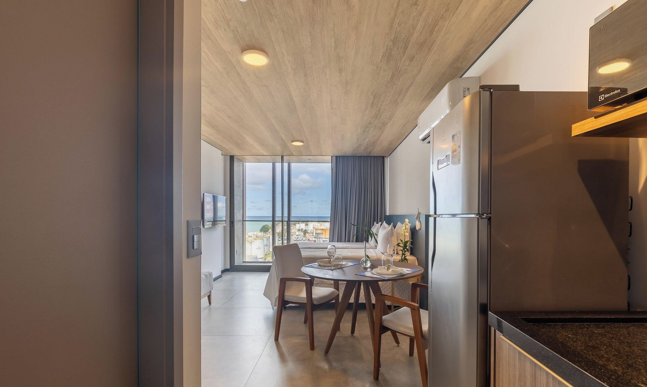 A modern kitchen with a built-in refrigerator, a dining area with a table and chairs, and a view of the sea through large windows.