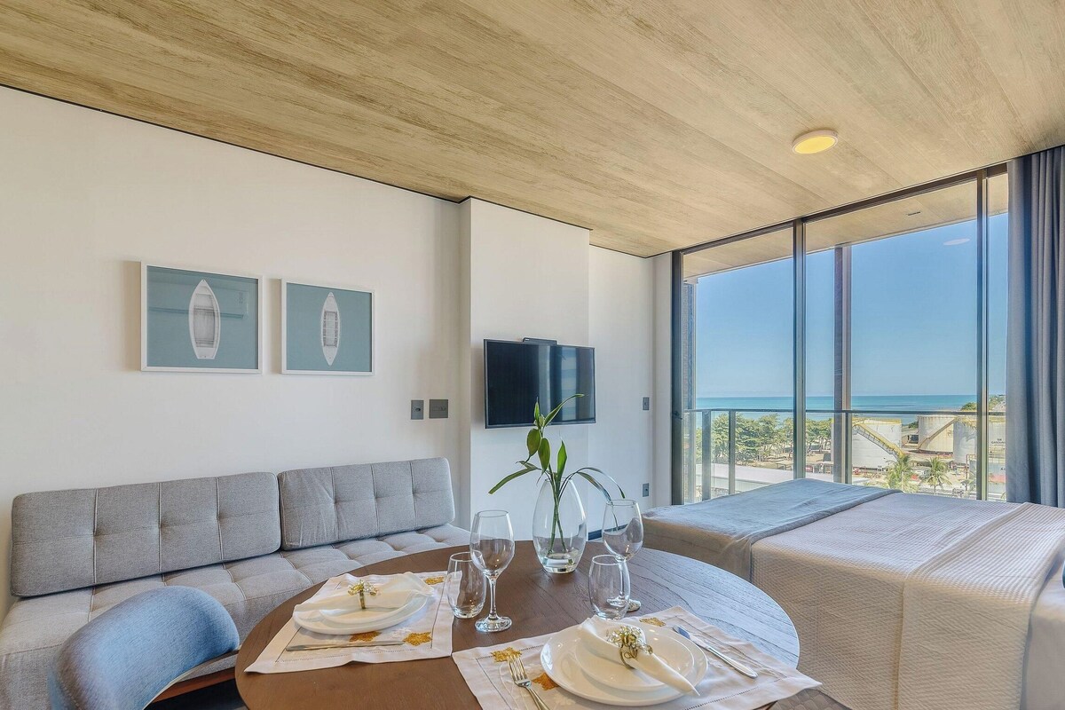 A modern hotel room with a dining area, a sofa, a TV, and a balcony with a view.
