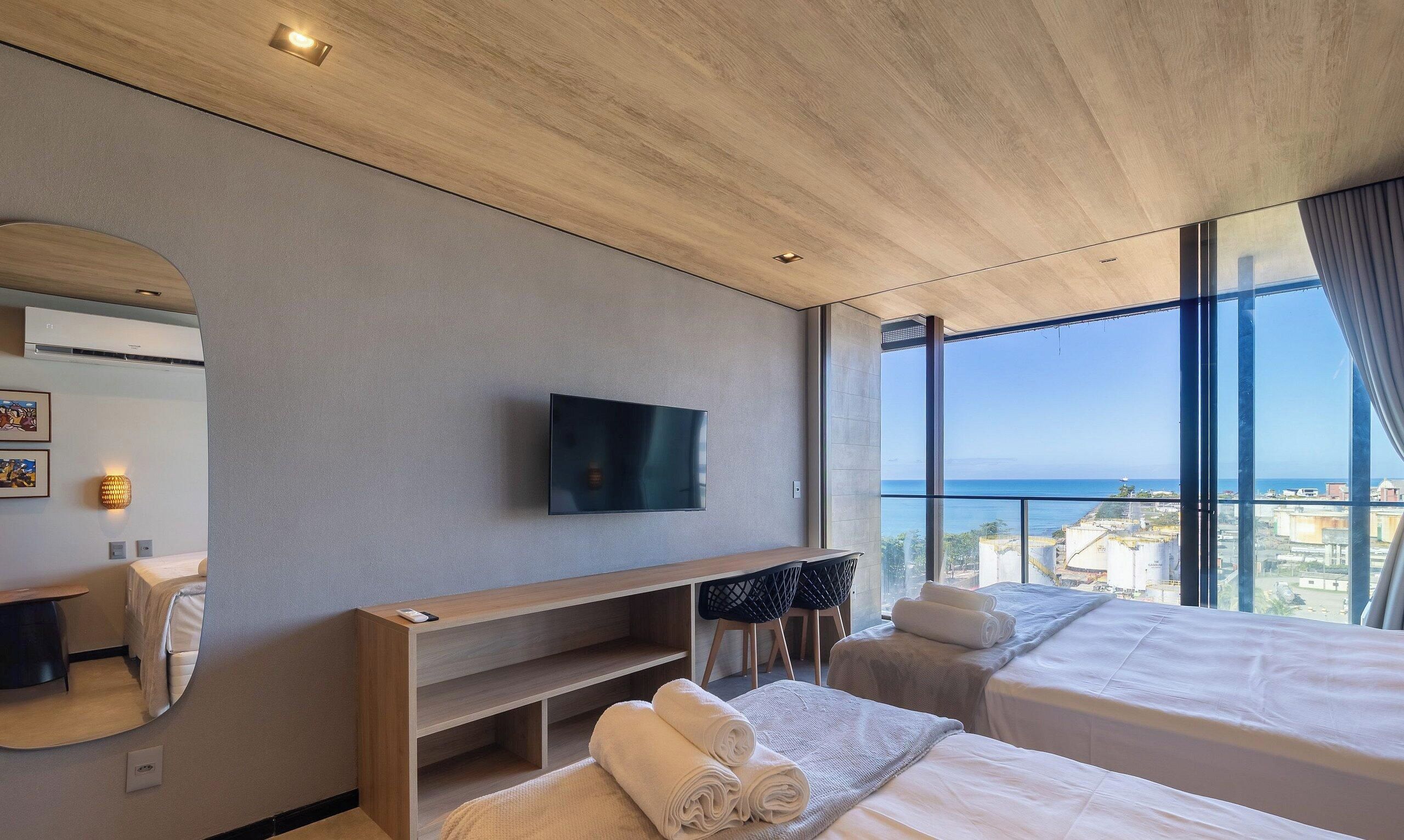 A modern hotel room with a large window, a flat-screen TV, a wooden desk, and a bed with towels.