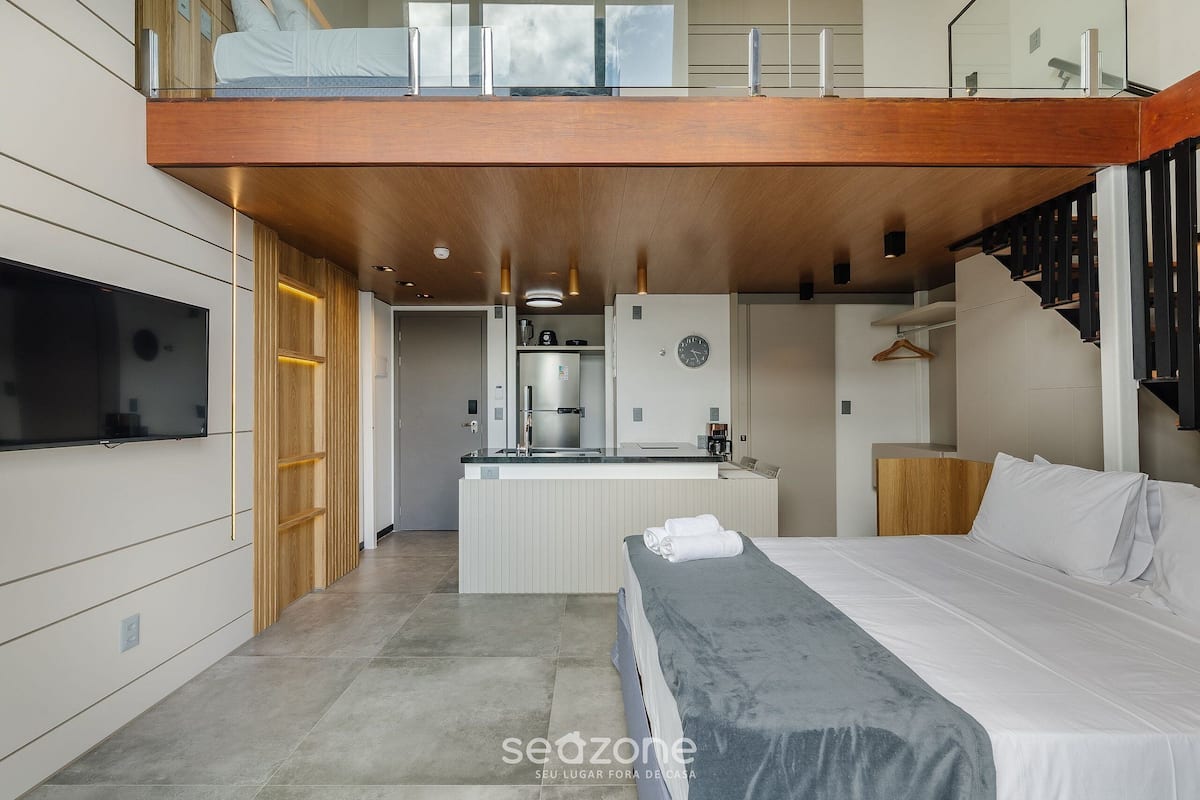 Una habitación de hotel moderna con una cama grande, una kitchenette pequeña, un televisor integrado y un balcón con barandilla de madera.