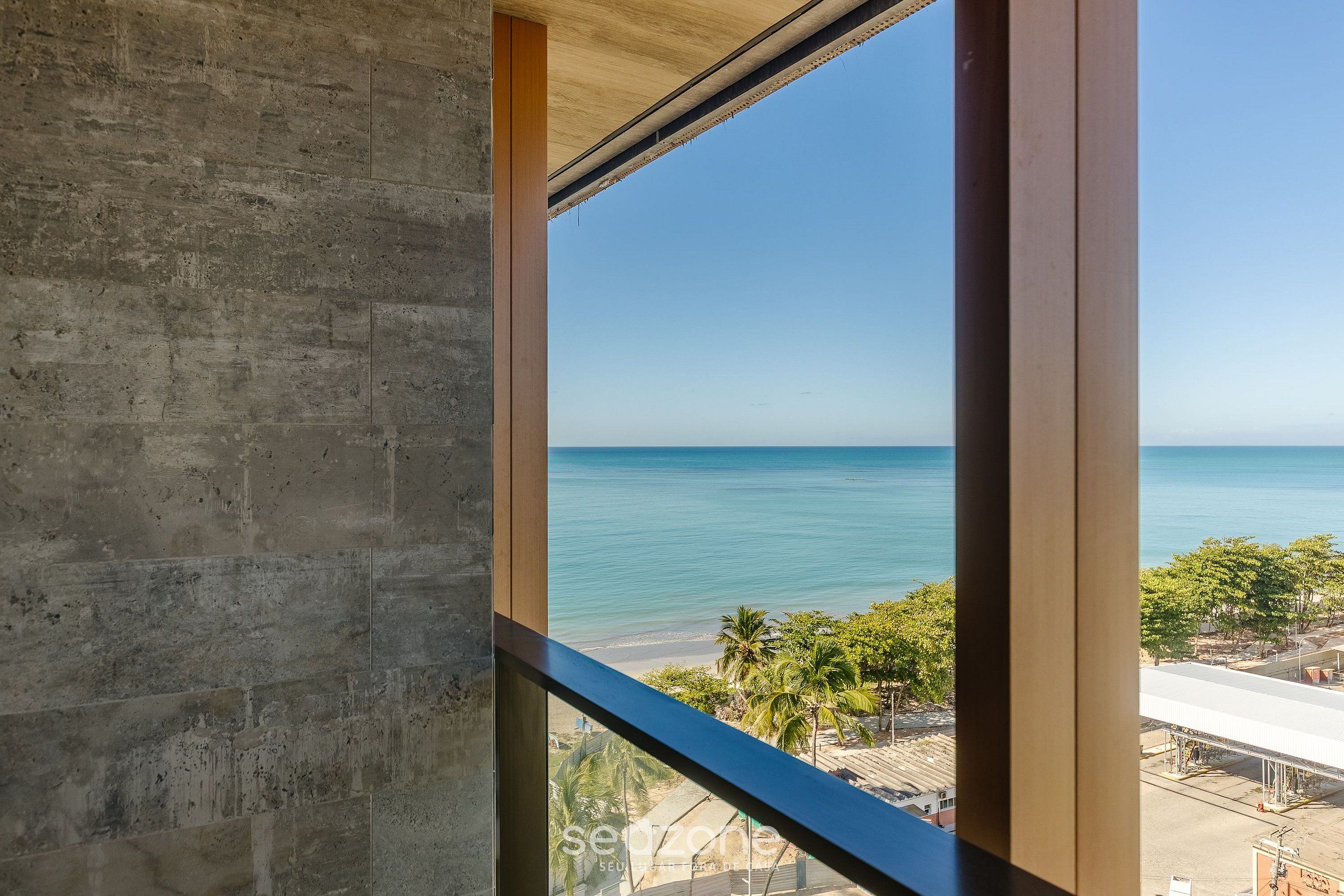 A view through a window with a concrete wall and wooden beams, overlooking a beach with palm trees and a clear sky.