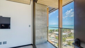 A room with a large window overlooking a coastal area, a flat-screen TV mounted on the wall, and a concrete pillar. - Nwts - Studios With Side sea View in Maceio III (Maceió)