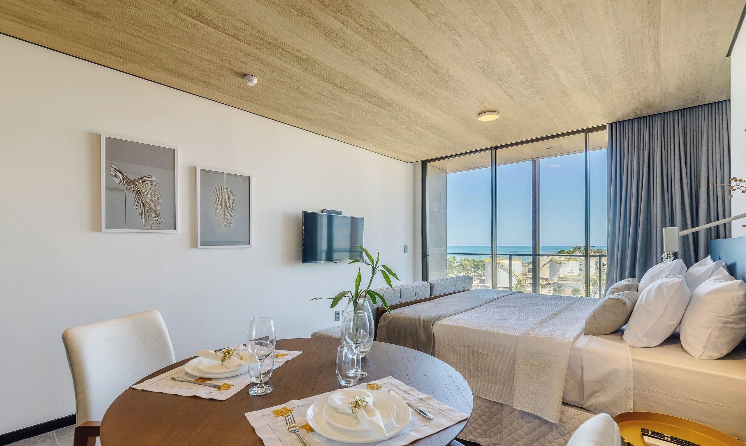 A hotel room with a dining area, a sofa, and a large window with a view.