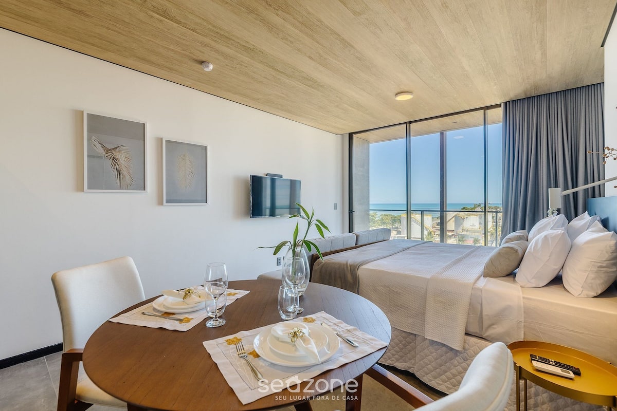 Habitación de hotel moderna con una mesa de comedor lista para dos personas, una cama con ropa de cama blanca y un ventanal con vistas a la ciudad.