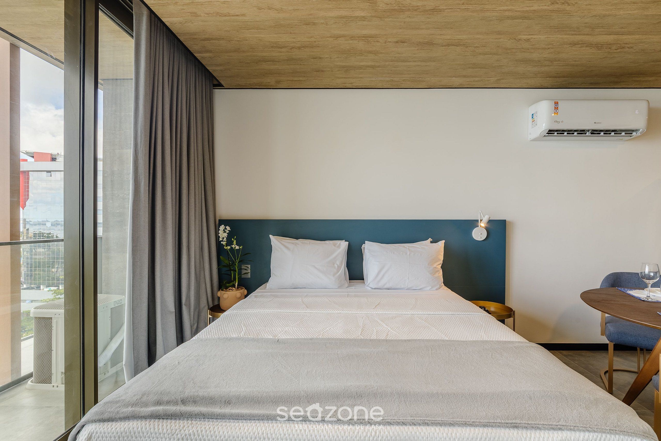 Habitación de hotel con una cama grande, una mesita de noche, una planta en maceta y vistas de la ciudad a través de un ventanal.