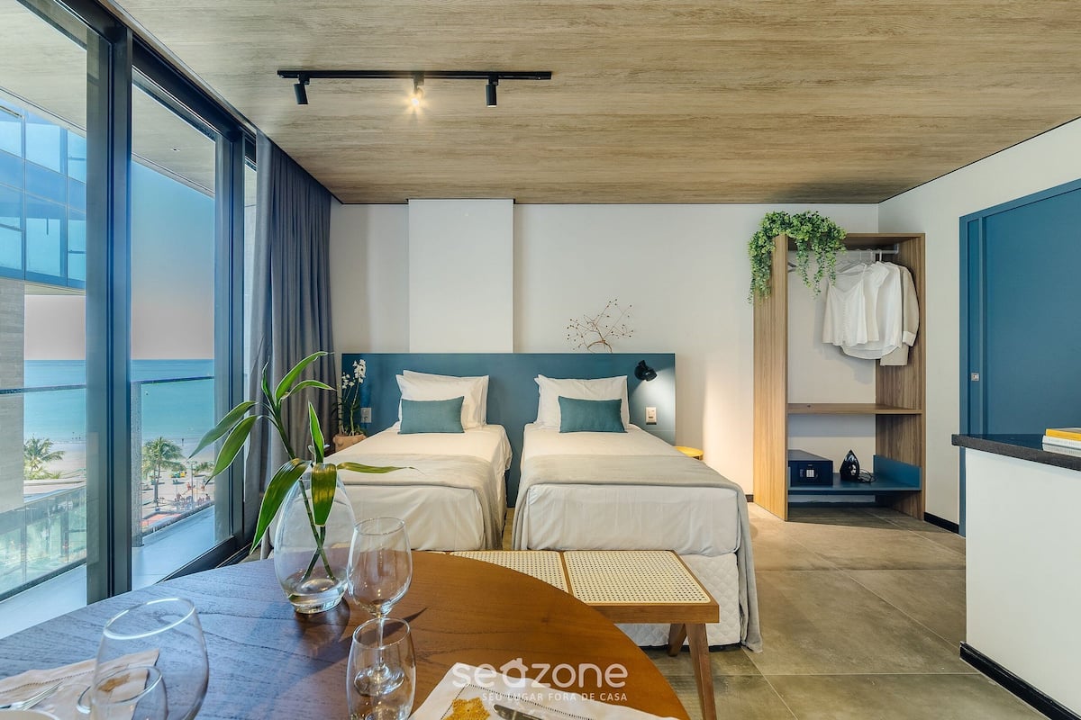 Habitación de hotel con dos camas, una mesa de madera con copas de vino, vista a la playa y una planta en la pared.
