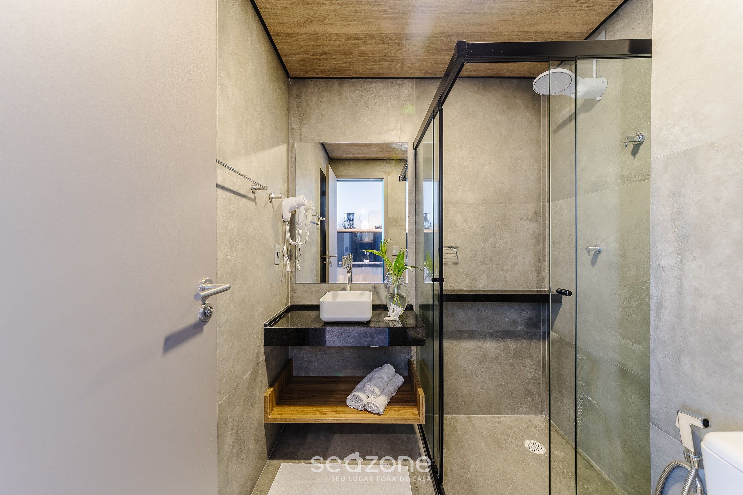A modern bathroom with a glass shower enclosure, a wooden shelf with towels, and a sink with a mirror above.