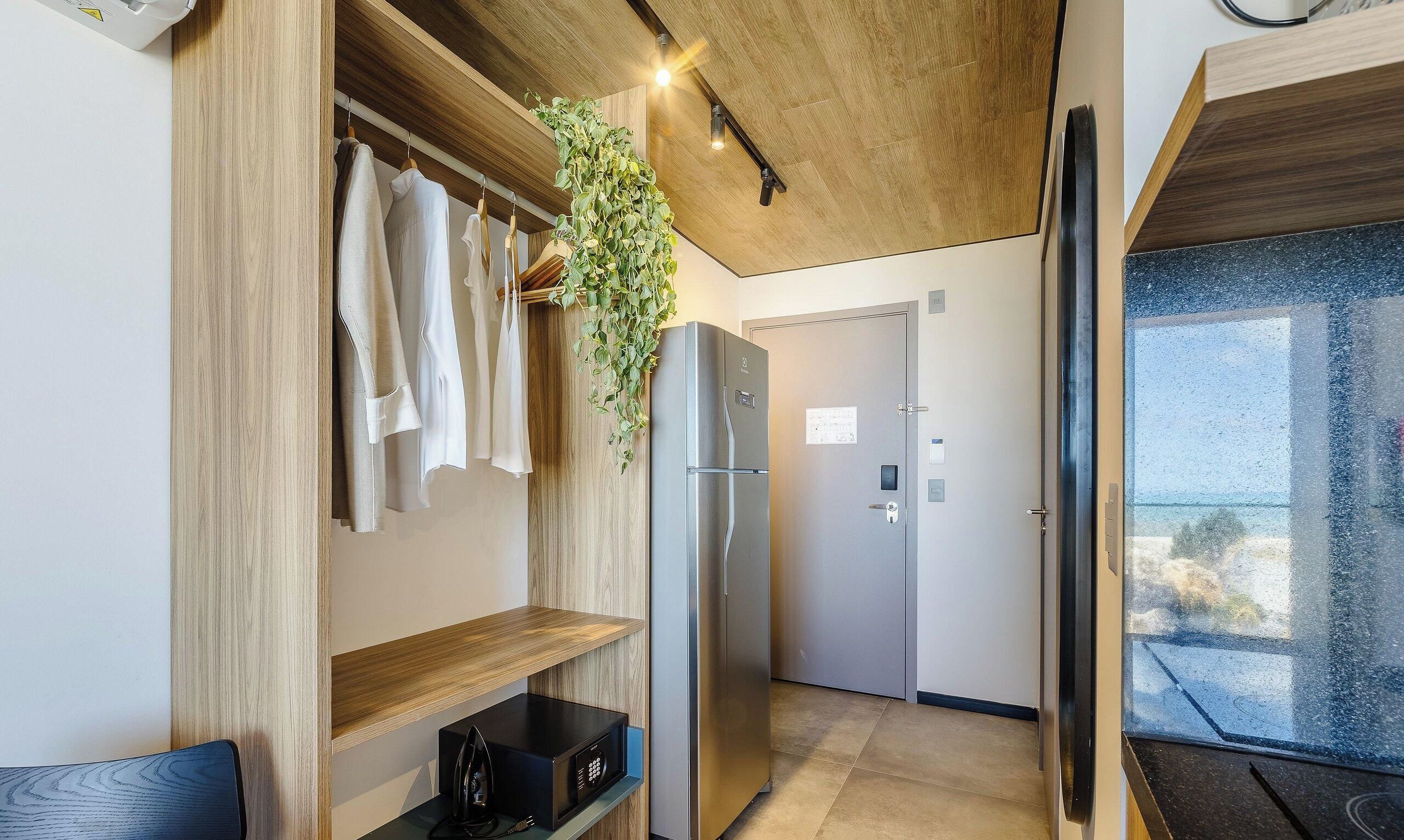 A modern hotel room with a wooden ceiling, a built-in wardrobe, a hanging plant, a refrigerator, and a view through a glass door.