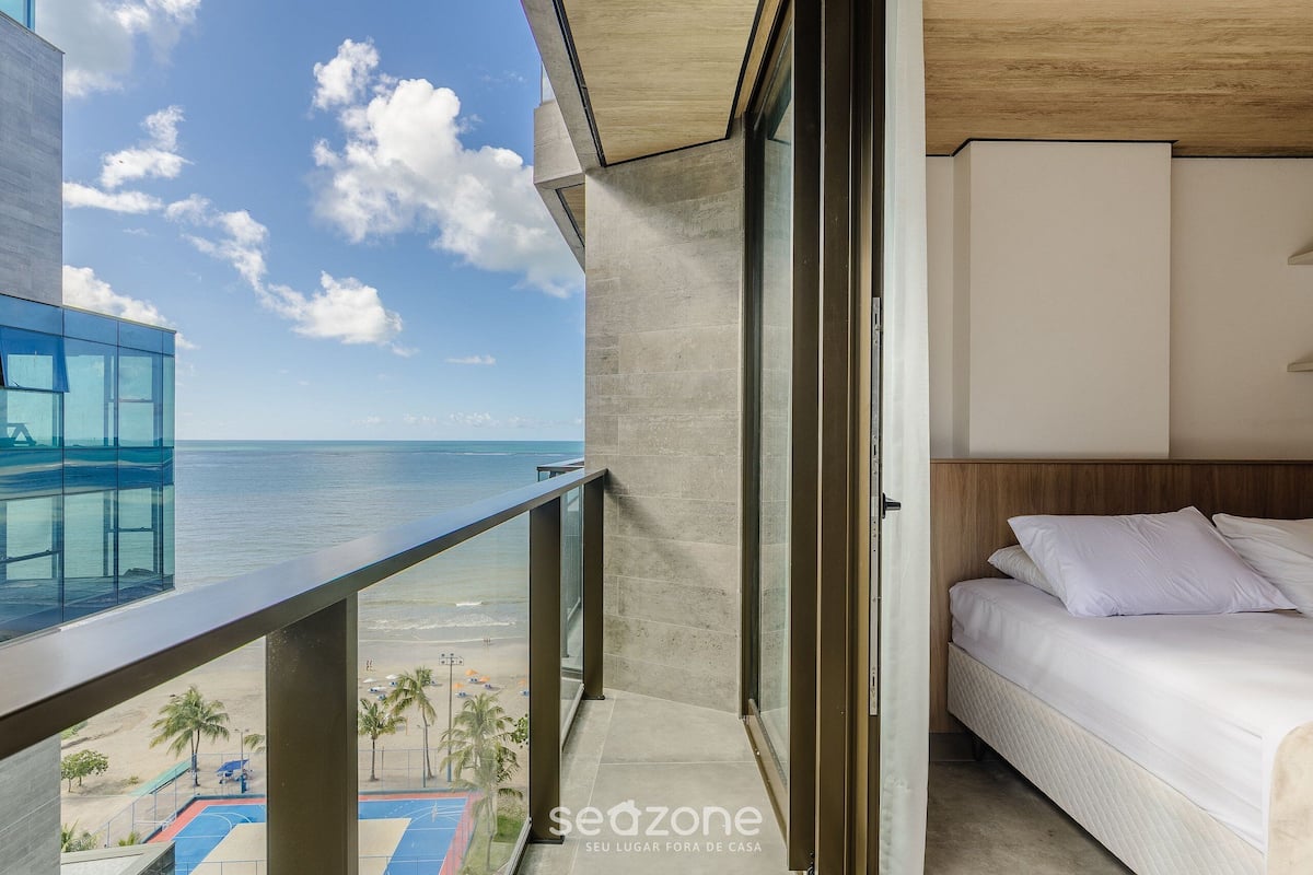 Un balcón con vista a la playa y el océano, una cama con ropa blanca y un edificio moderno con ventanas de vidrio.