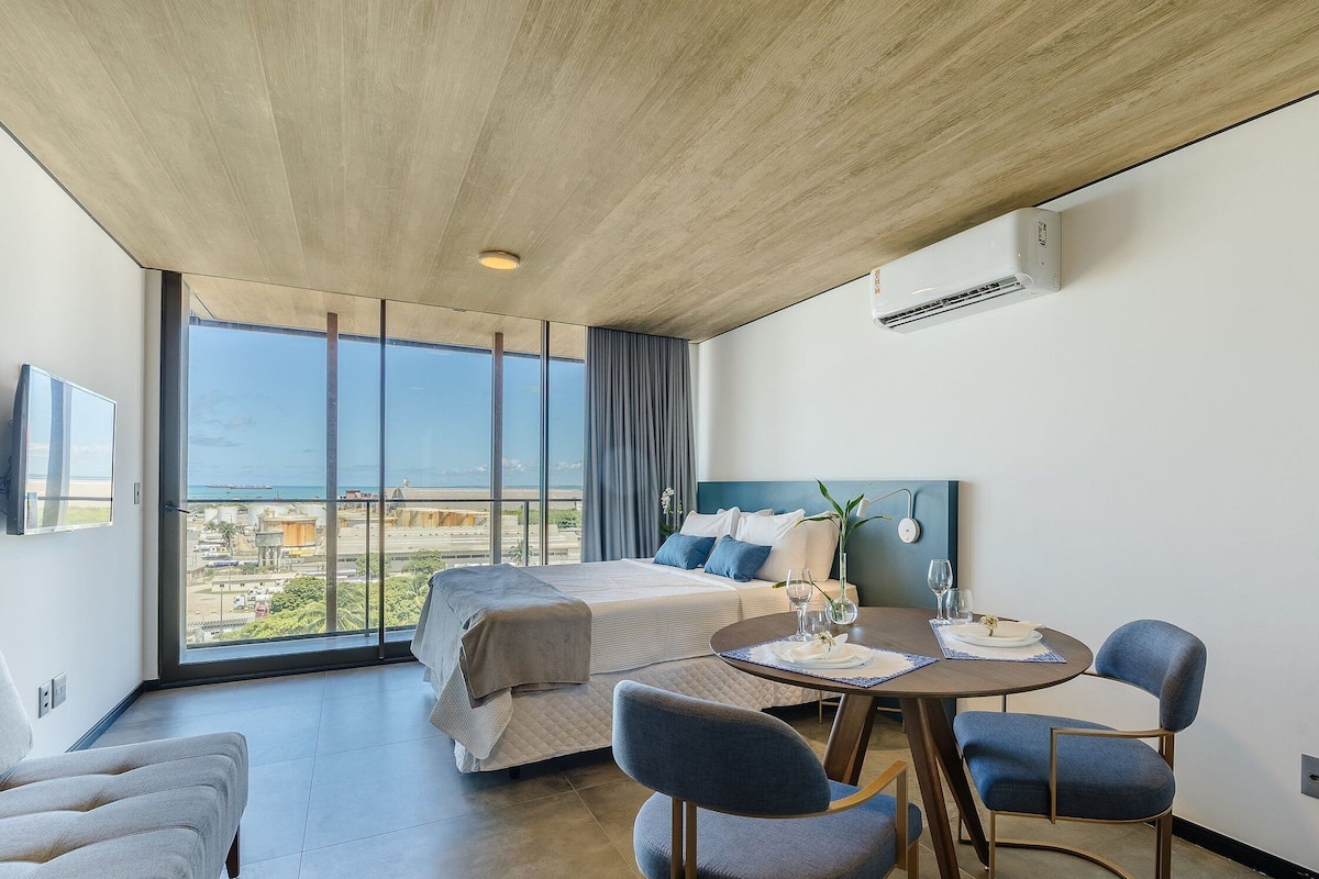 Una habitación de hotel moderna con una cama grande, una mesa de comedor con sillas, vistas a la ciudad y un equipo de aire acondicionado.