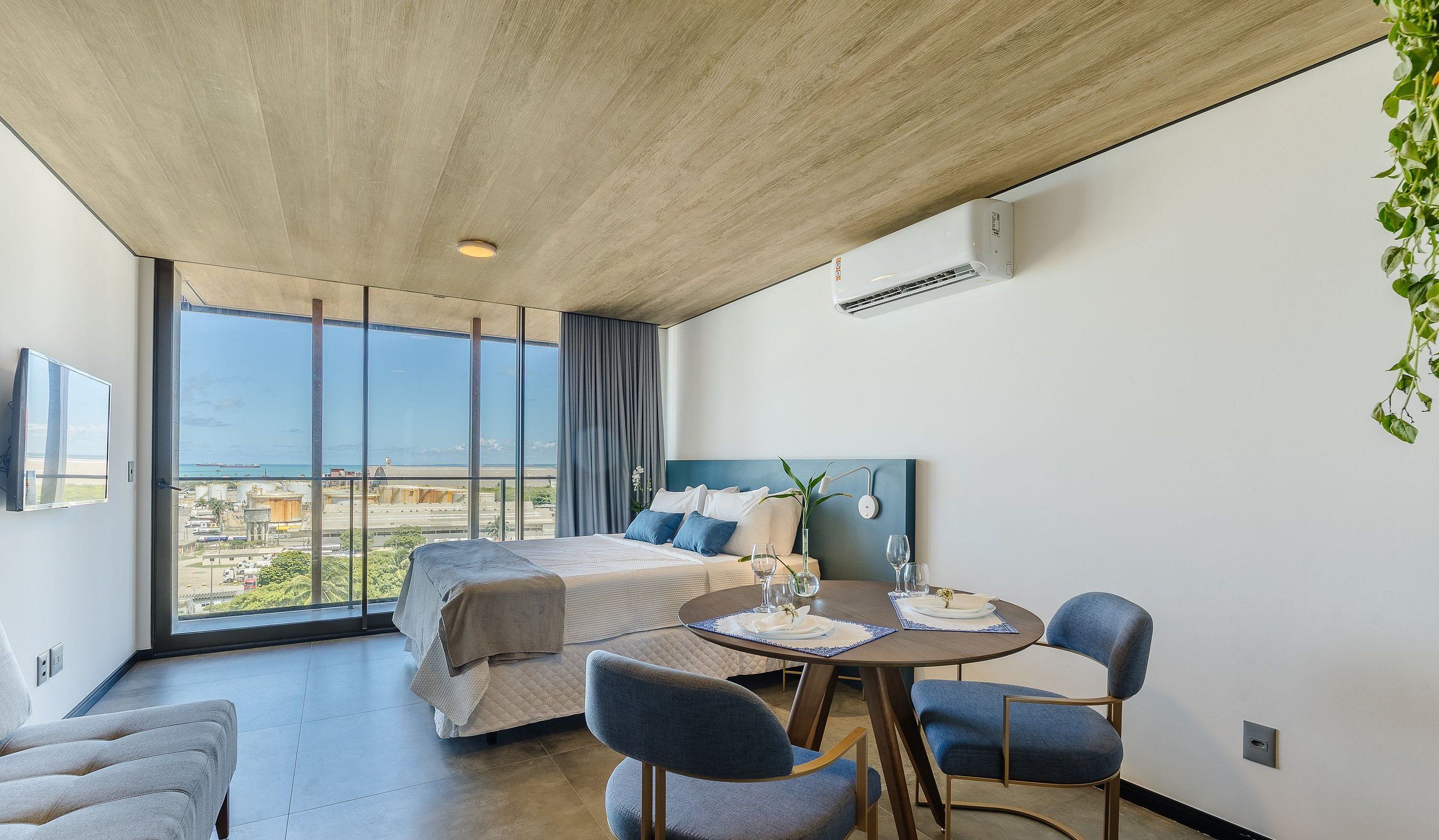 A modern hotel room with a large bed, a dining table with chairs, a view of the city, and an air conditioning unit.