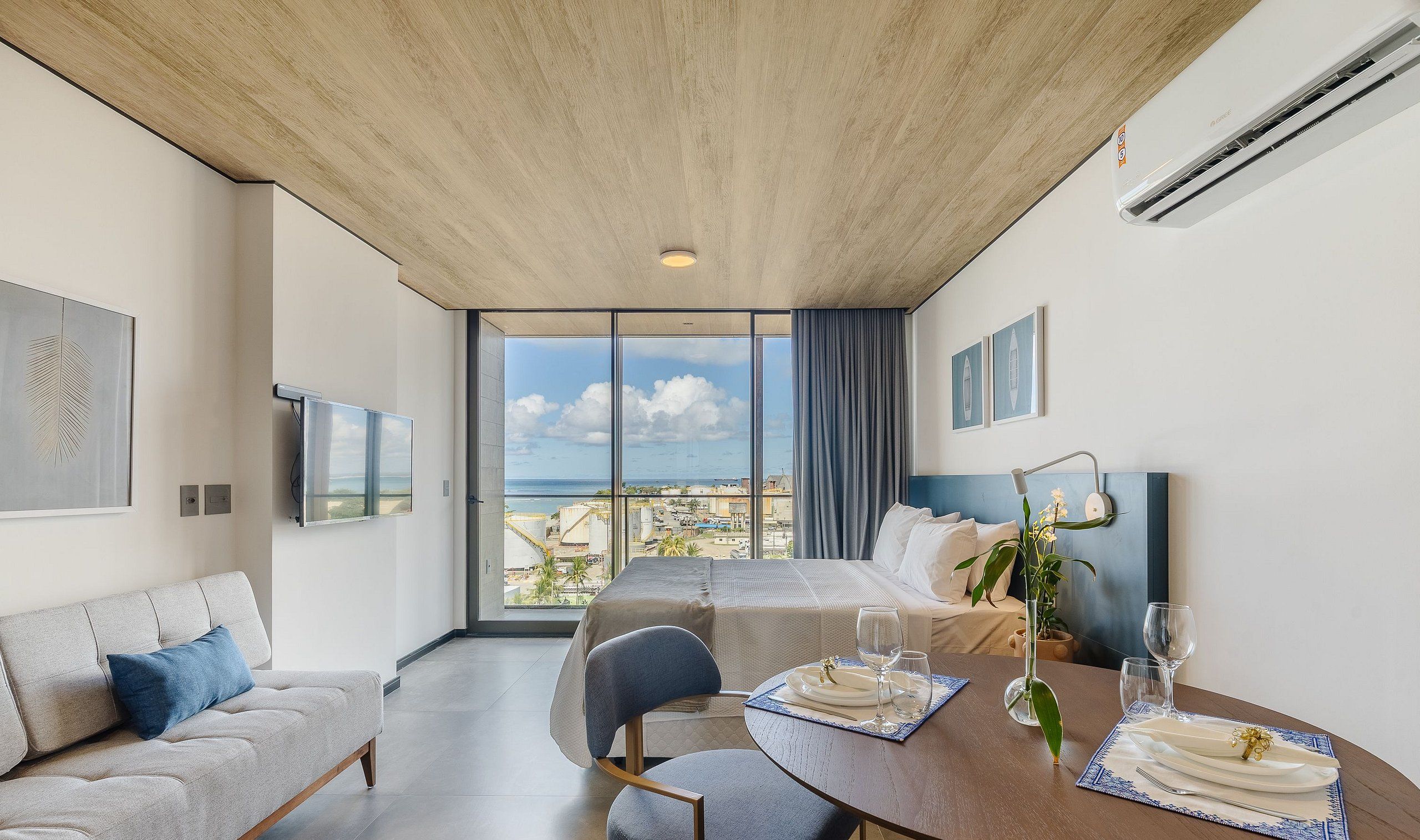 A modern hotel room with a large window, a dining table set for two, a sofa, and a bed.