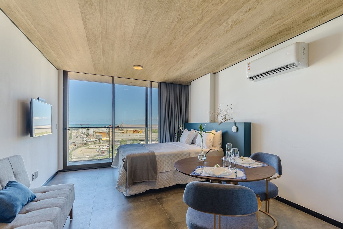 Una habitación de hotel moderna con un ventanal grande, una cama, una mesa de comedor con sillas y un equipo de aire acondicionado.