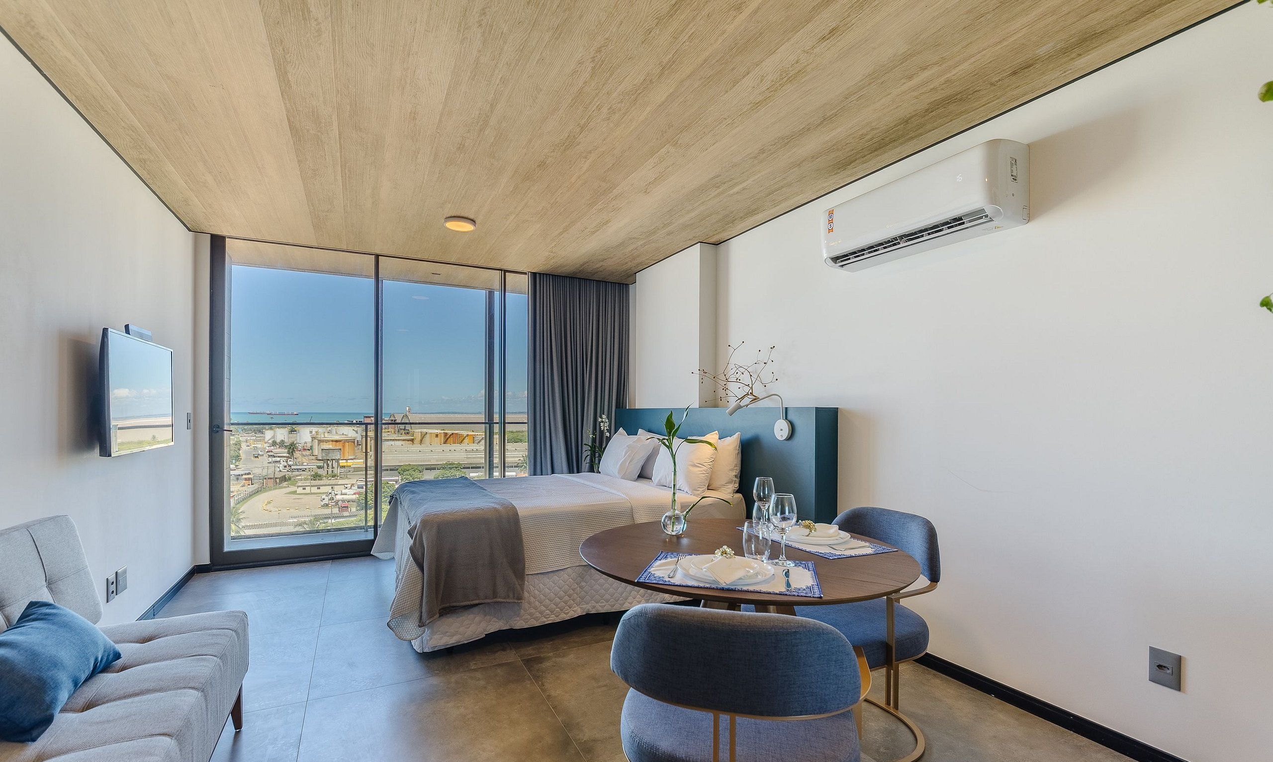 Una habitación de hotel moderna con un ventanal grande, una cama, una mesa de comedor con sillas y un equipo de aire acondicionado.