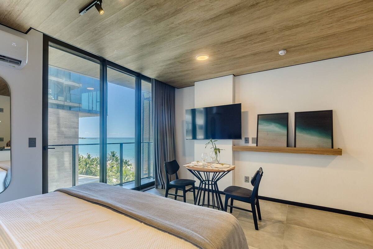 Una habitación de hotel moderna con una cama grande, un balcón con vista al mar, un pequeño comedor y un televisor en la pared.