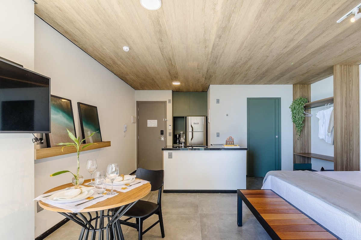Una habitación de hotel moderna con techo de madera, un área de comedor con mesa para dos, una kitchenette y una cama.