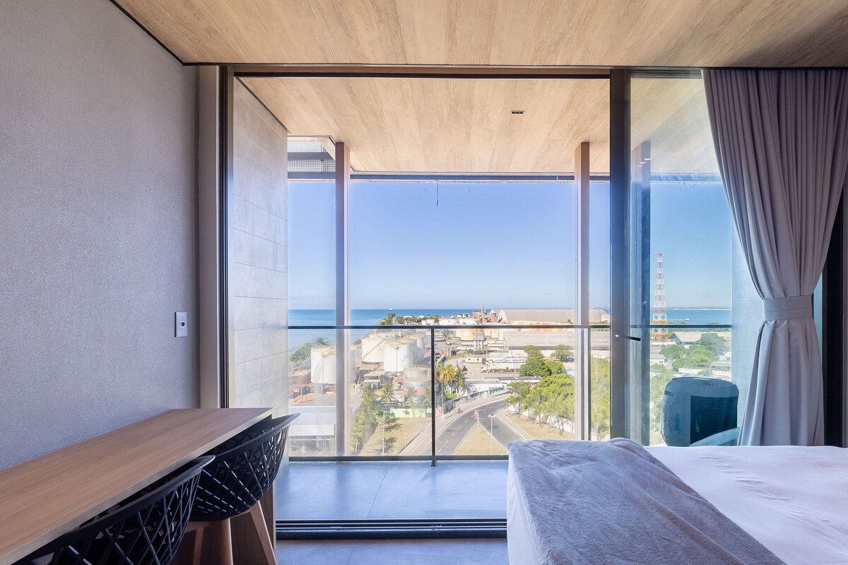 Een moderne hotelkamer met een ruim balkon, een bed, een bureau en uitzicht op de stad en zee.
