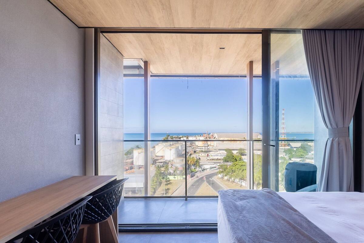 Habitación de hotel moderna con un balcón amplio, una cama, un escritorio y vistas a la ciudad y al mar.