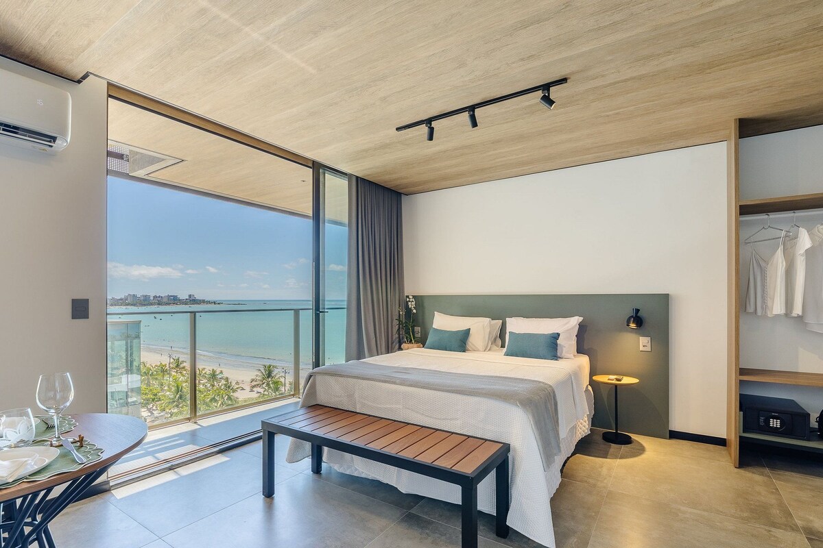Una habitación de hotel moderna con una cama grande, una mesita de noche, un banco, una pequeña mesa con una copa de vino y vista a la playa.