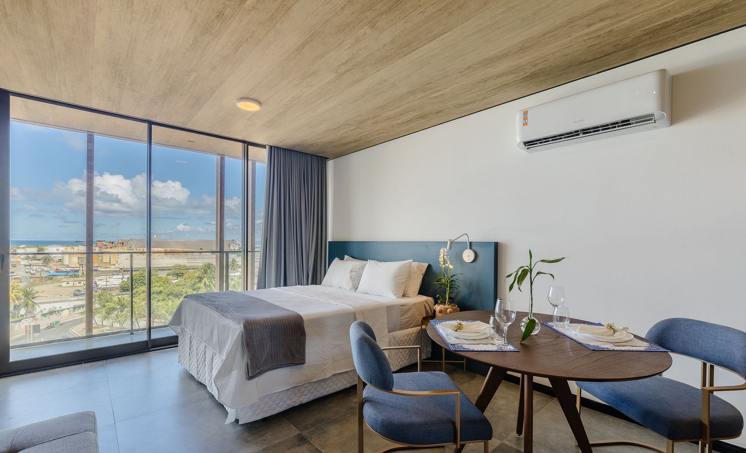 Quarto de hotel moderno com uma cama grande, uma mesa de jantar com cadeiras e vista para a cidade através de amplas janelas.