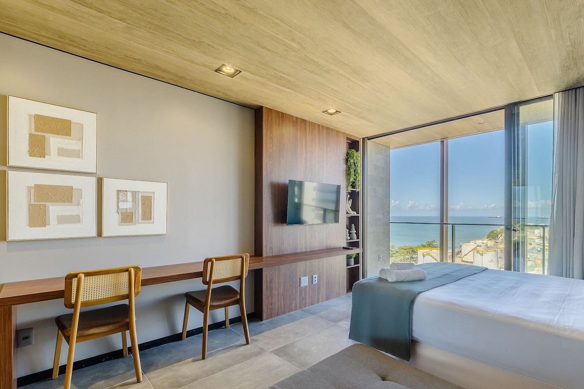 Una habitación de hotel moderna con una cama grande, un escritorio con una silla, un televisor de pantalla plana en la pared y un balcón con vistas al mar.