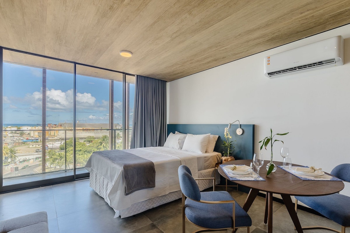 Una habitación de hotel moderna con una cama grande, una mesa de comedor con sillas y vistas de la ciudad a través de amplios ventanales.